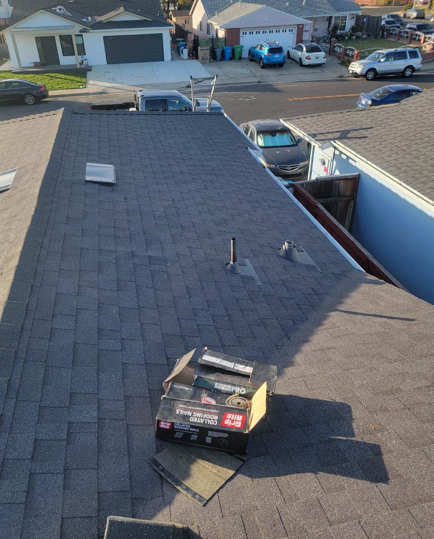 A freshly completed dark grey architectural shingle roof replacement by New Concept Roofing Inc., showing clean line installations, vent pipes, and a box of roofing nails on the surface.